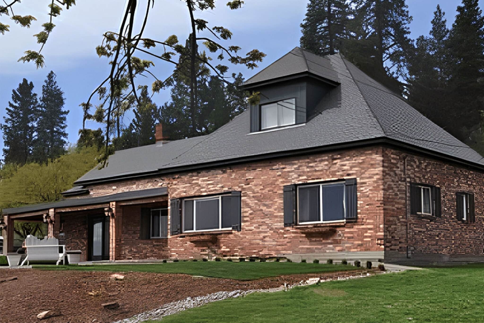 A brick house with a dark, steep roof and several large windows sits on a grassy lawn, surrounded by trees under a clear sky. Welcome to Farmgirl Gourmet—relax on the covered patio with outdoor furniture visible on the left side.