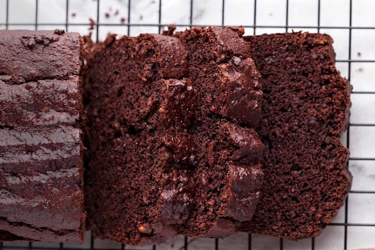 Three slices of rich, moist chocolate zucchini bread rest on a black cooling rack, showing a dark, fudgy texture and a shiny, slightly cracked top—a delicious result from an easy chocolate zucchini bread recipe.