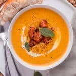 A bowl of roasted butternut squash soup topped with sausage, cream, and sage leaves, served with a piece of crusty bread.