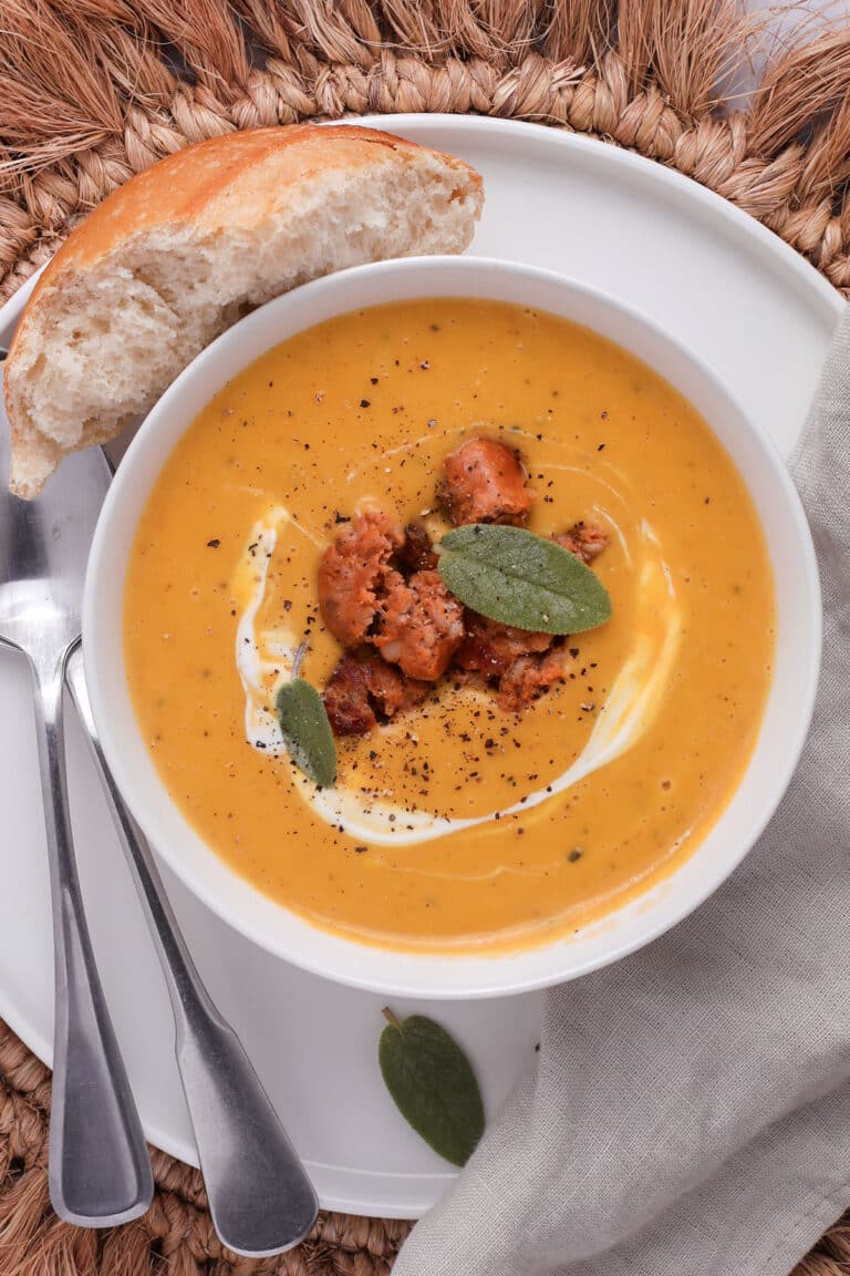 A bowl of roasted butternut squash soup topped with sausage, cream, and sage leaves, served with a piece of crusty bread.
