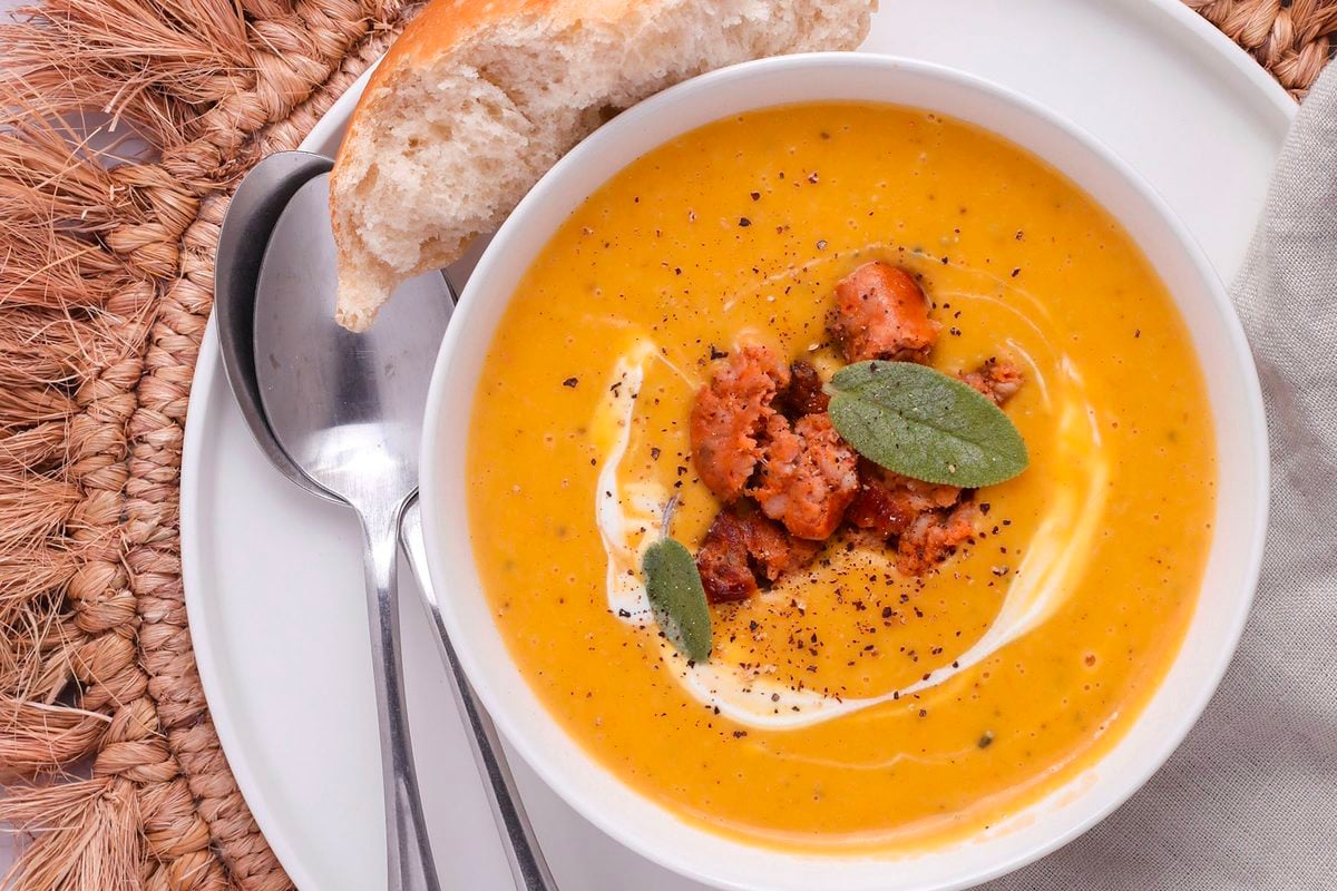 A bowl of creamy roasted butternut squash soup topped with sausage, sage leaves, and a swirl of cream, served with a piece of crusty bread and a spoon.
