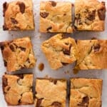 Nine blondie bars with chunks of rolo candies are arranged on a sheet of crumpled parchment paper. The bars are golden brown with visible swirls and pieces of chocolate throughout.