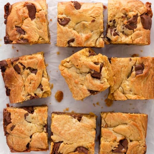 Nine blondie bars with chunks of rolo candies are arranged on a sheet of crumpled parchment paper. The bars are golden brown with visible swirls and pieces of chocolate throughout.