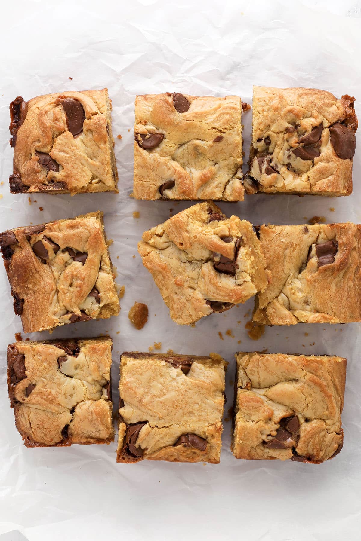 Nine blondie bars with chunks of rolo candies are arranged on a sheet of crumpled parchment paper. The bars are golden brown with visible swirls and pieces of chocolate throughout.