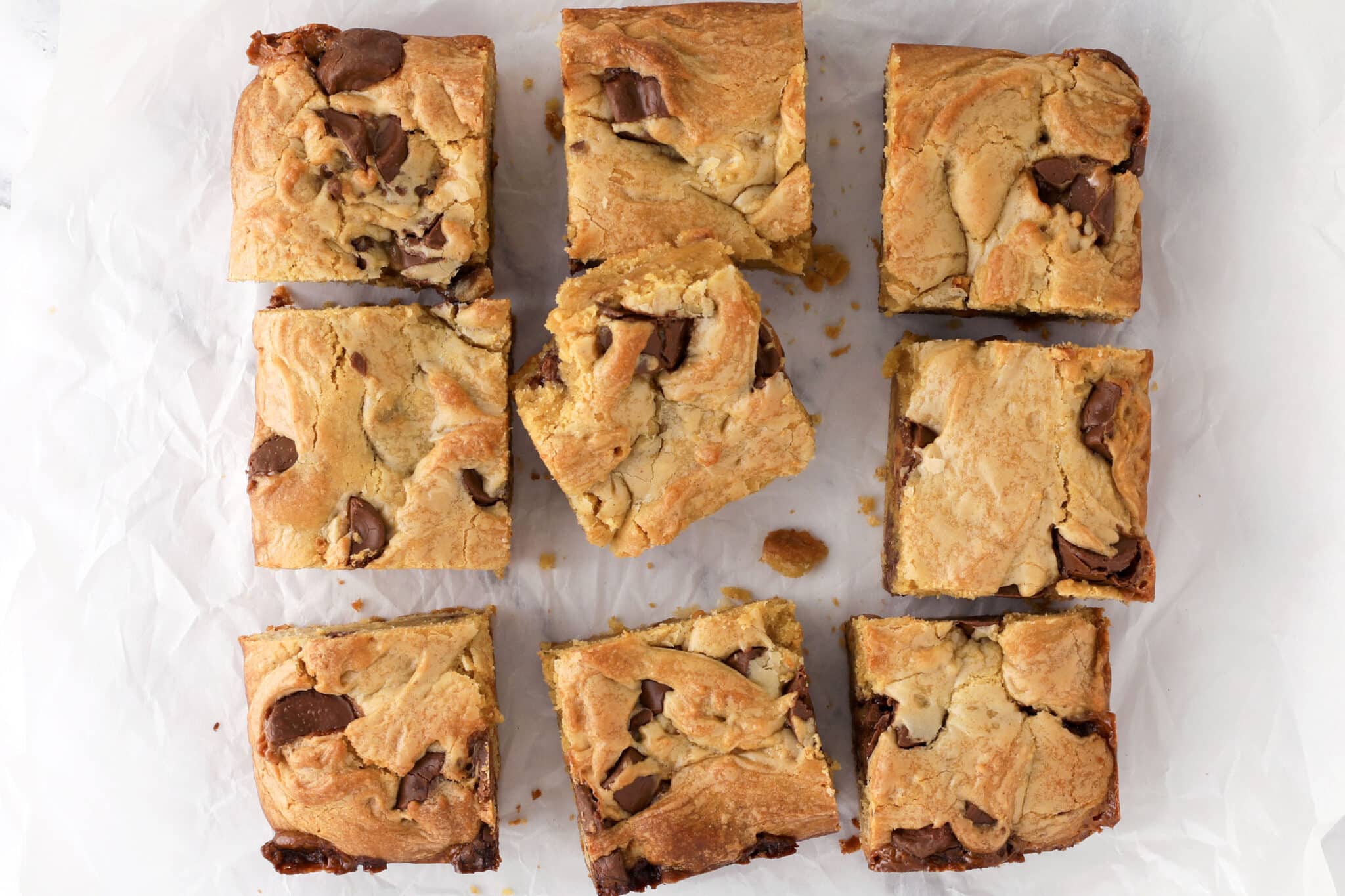 Nine squares of gooey blondies with chocolate chunks are arranged on white parchment paper, viewed from above. The blondies are golden brown with visible swirls and pieces of melted chocolate.