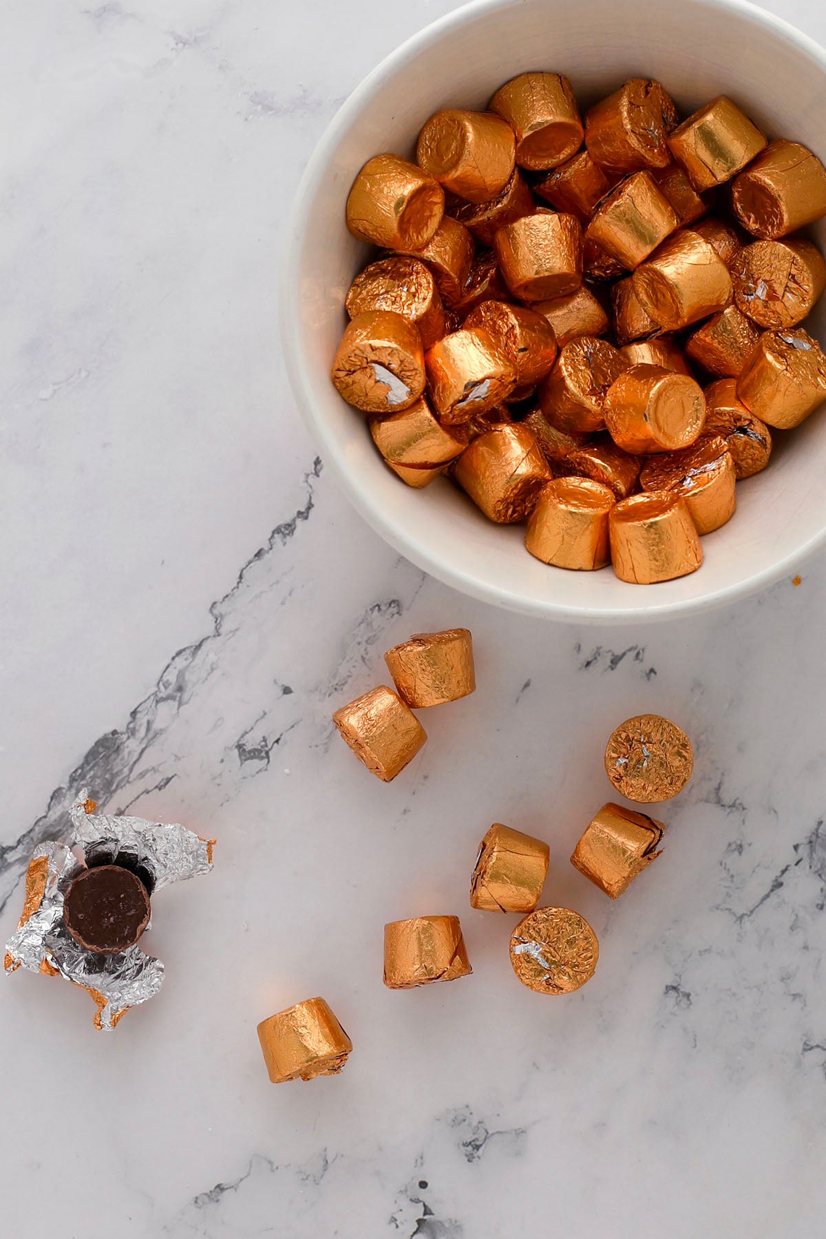 A bowl filled with gold-wrapped chocolate candies sits on a marble surface, with several unwrapped chocolates scattered nearby and one partially unwrapped to reveal the dark center.