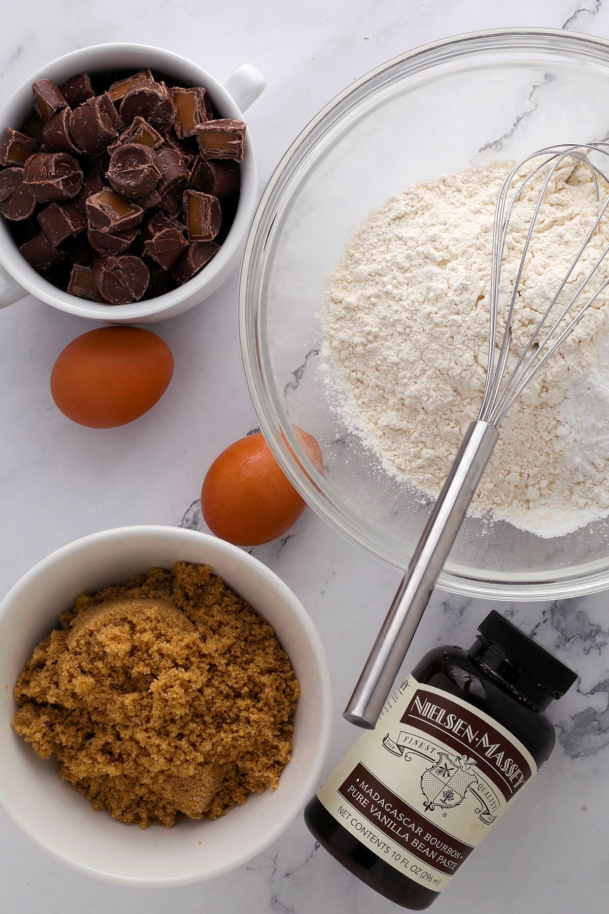 Baking ingredients on a white surface: a bowl of flour with a whisk, two brown eggs, a bowl of brown sugar, a cup of chopped chocolate, and a bottle of vanilla extract.