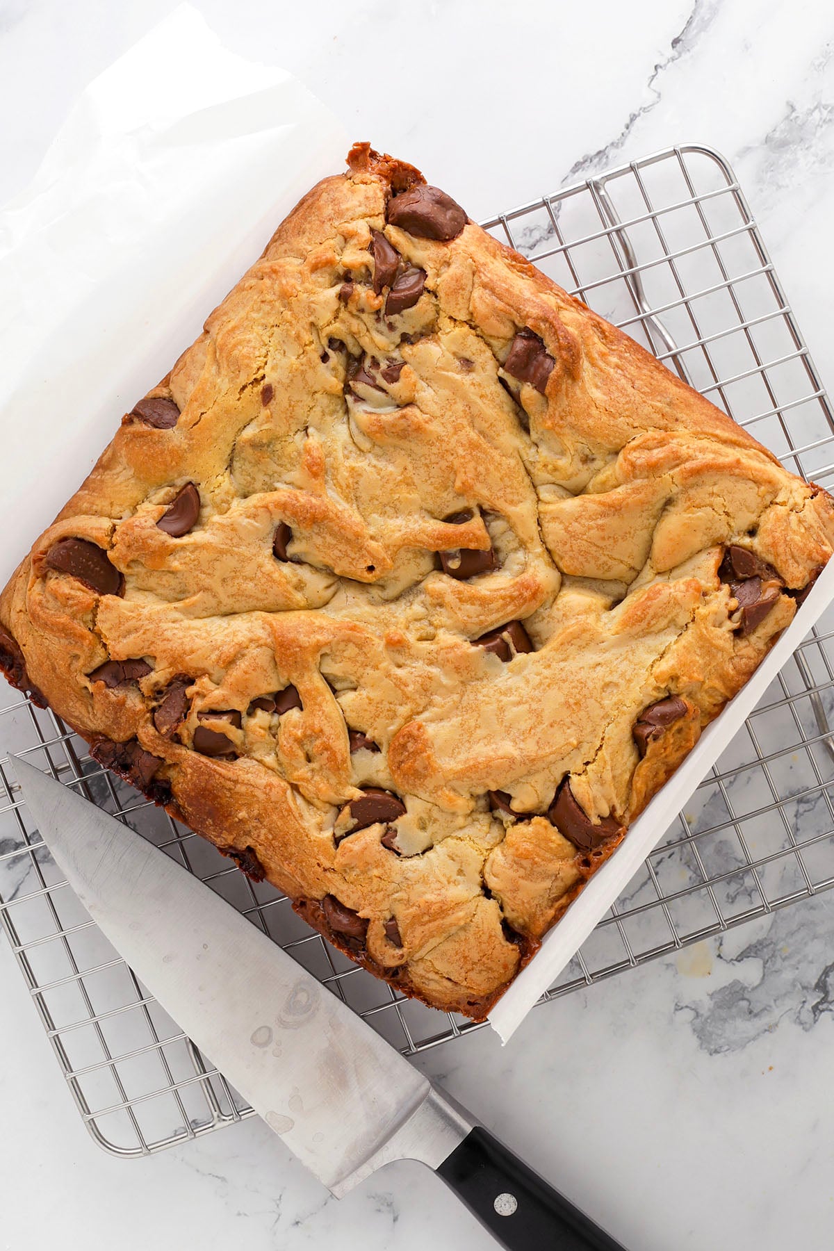 A square pan of golden-brown, freshly baked blondies with melted chocolate chunks sits on a wire cooling rack atop a marble surface. A knife rests next to the pan.