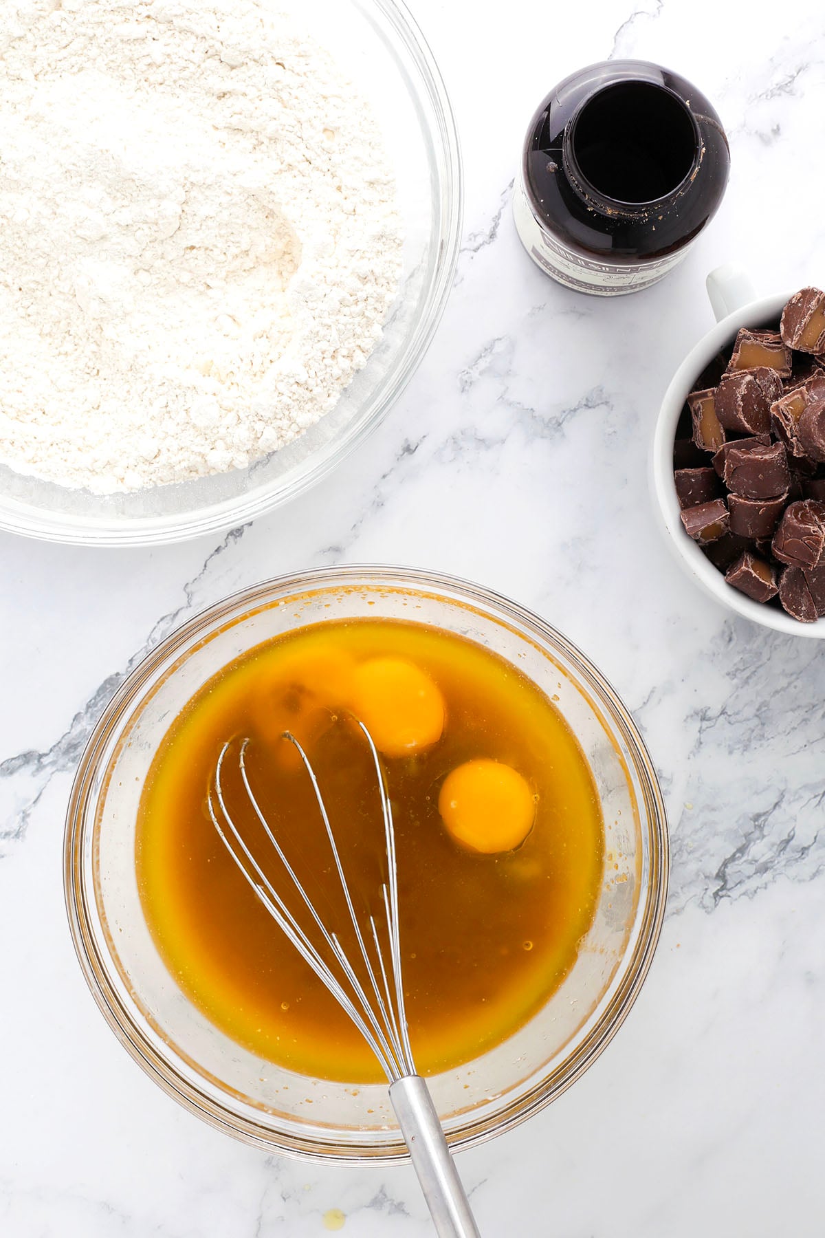 A bowl with eggs, sugar, and melted butter being whisked sits on a marble counter next to a bowl of flour, a cup of chopped chocolate, and a bottle of vanilla extract.