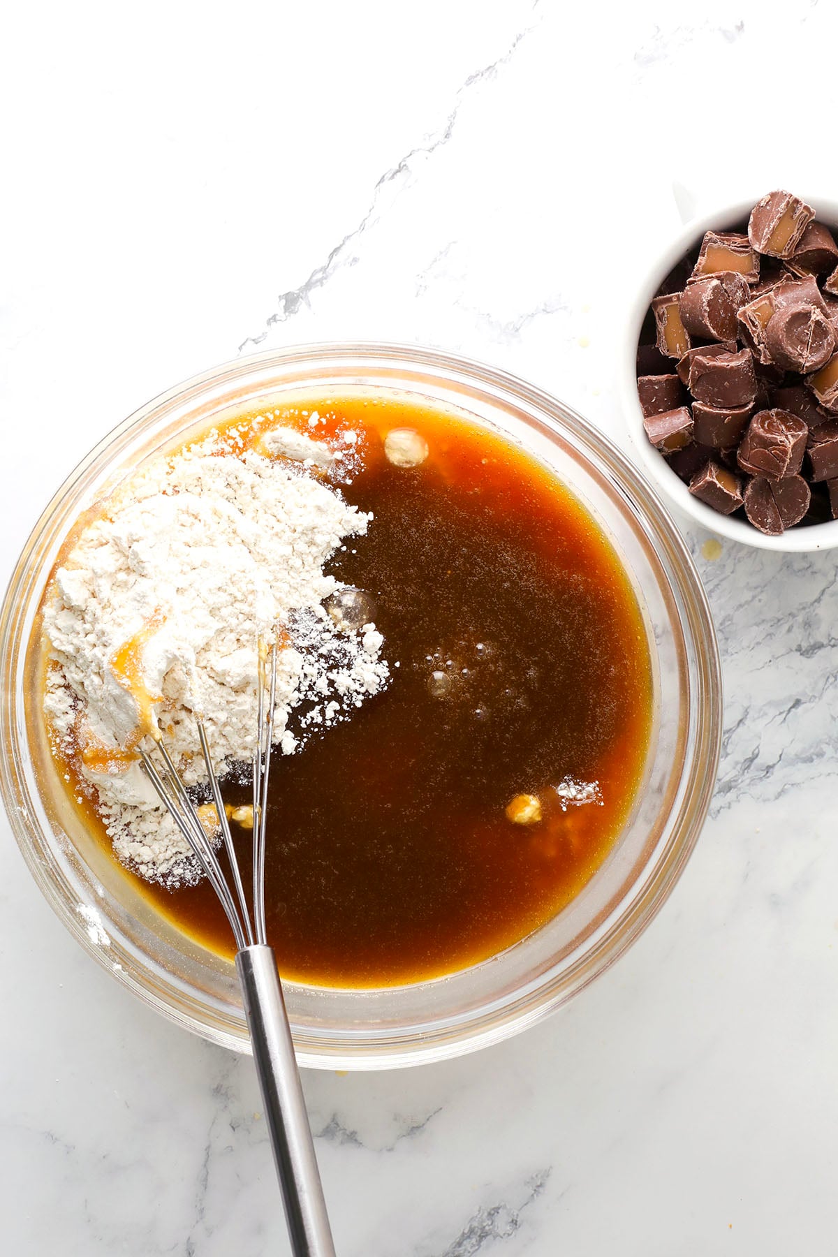 A glass mixing bowl with flour, brown liquid, and a whisk on a white marble surface; a white bowl filled with chocolate chunks is nearby.