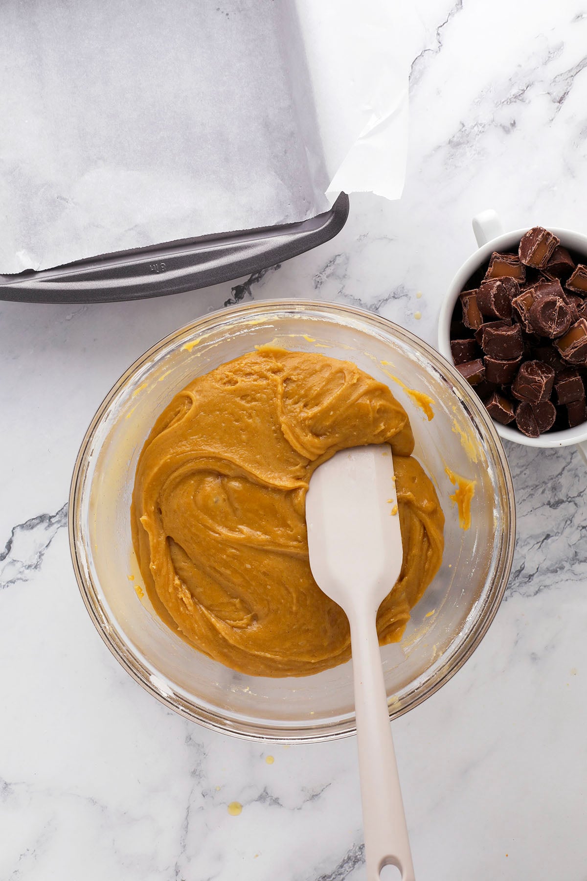A glass bowl filled with light brown batter is being mixed with a spatula on a marble surface. Nearby, there is a baking pan lined with parchment paper and a white cup filled with chocolate candies.