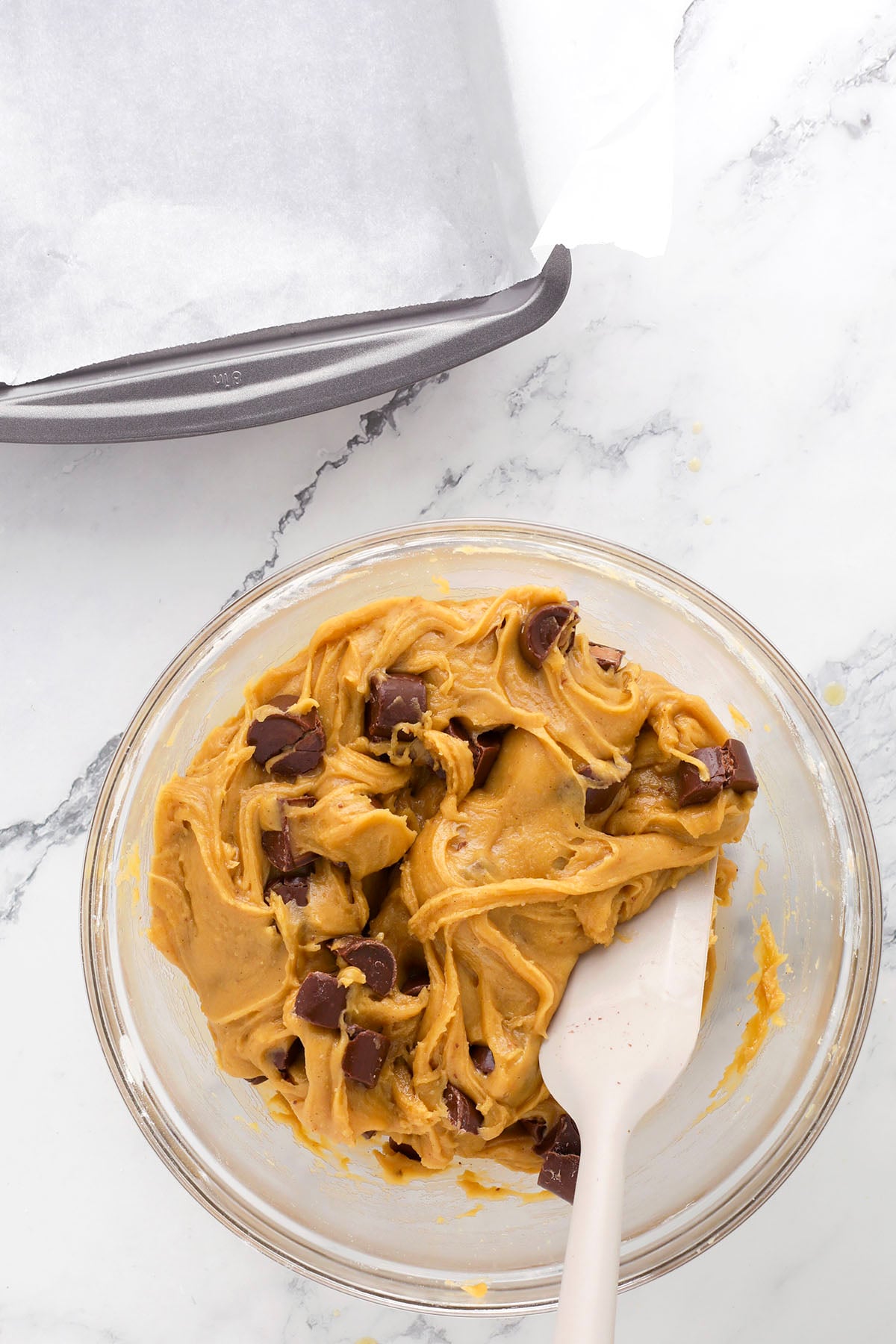 A glass bowl filled with chocolate chip cookie dough and a white spatula, sitting on a marble countertop next to a parchment-lined baking pan.