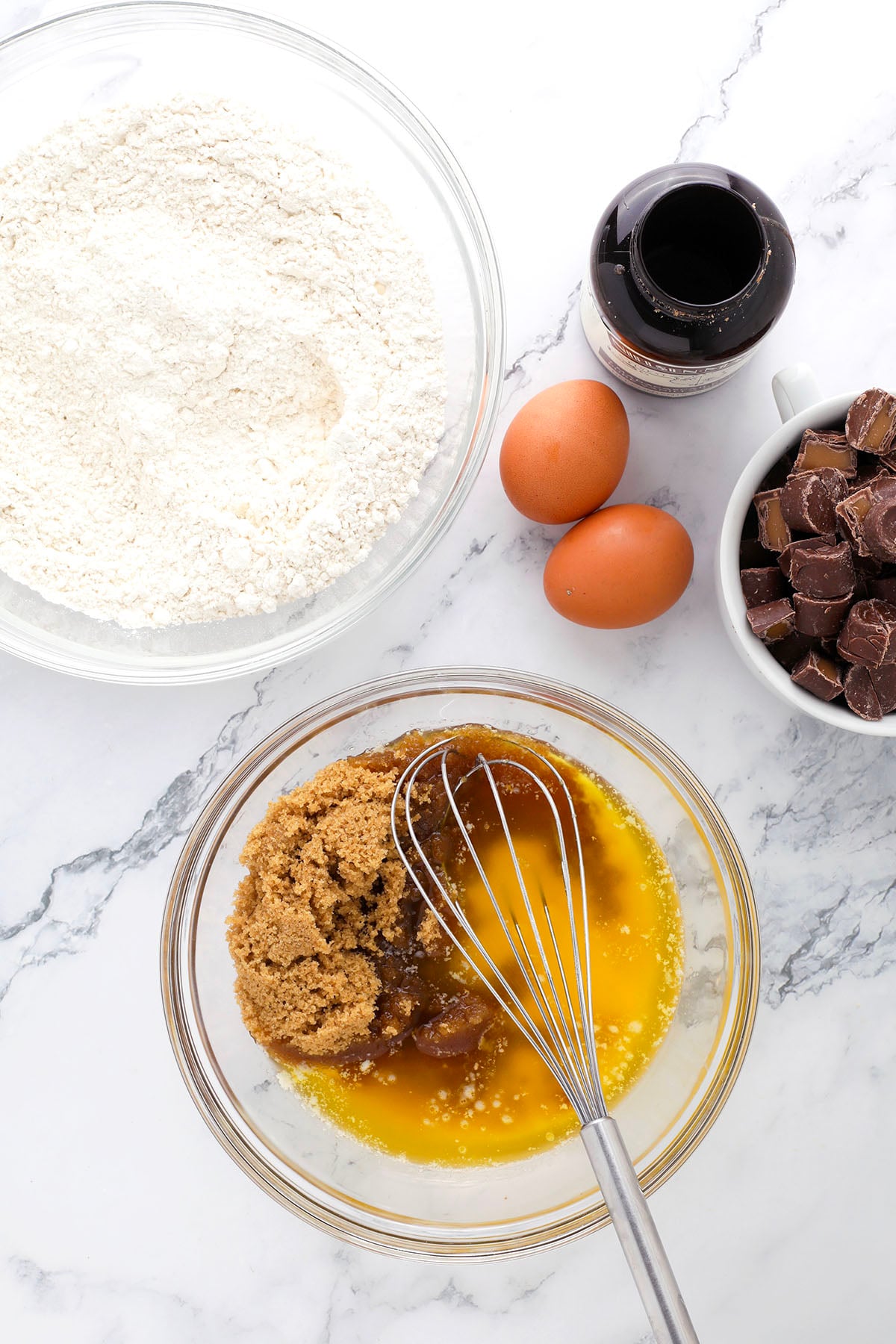 A whisk rests in a bowl of melted butter and brown sugar. Nearby are a bowl of flour, two eggs, a bottle of vanilla, and a cup of chocolate chunks on a marble countertop.