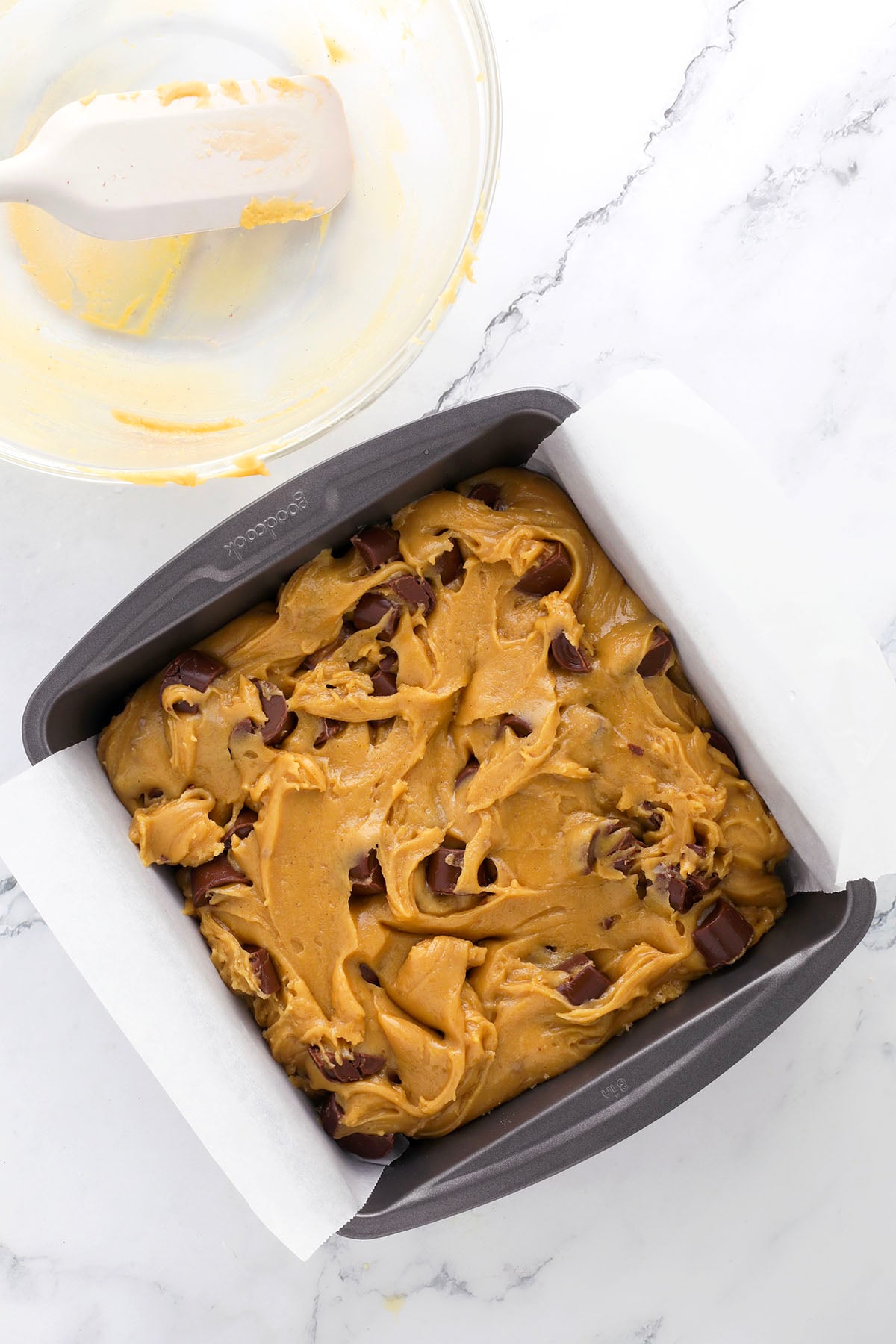 A square baking pan lined with parchment paper is filled with unbaked cookie dough mixed with chocolate chunks. A glass bowl with leftover dough and a spatula sits nearby on a marble countertop.
