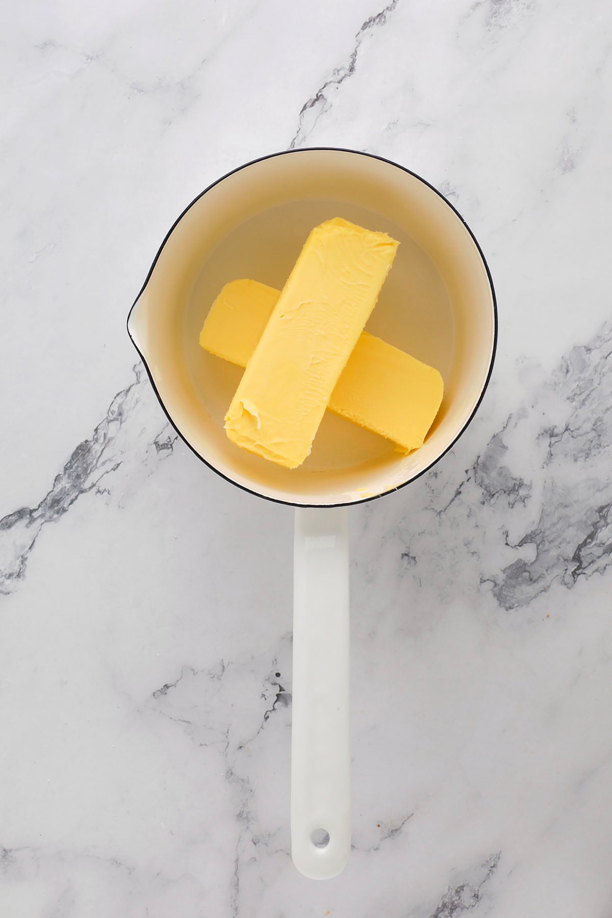A white saucepan with two sticks of butter inside, placed on a white marble surface.