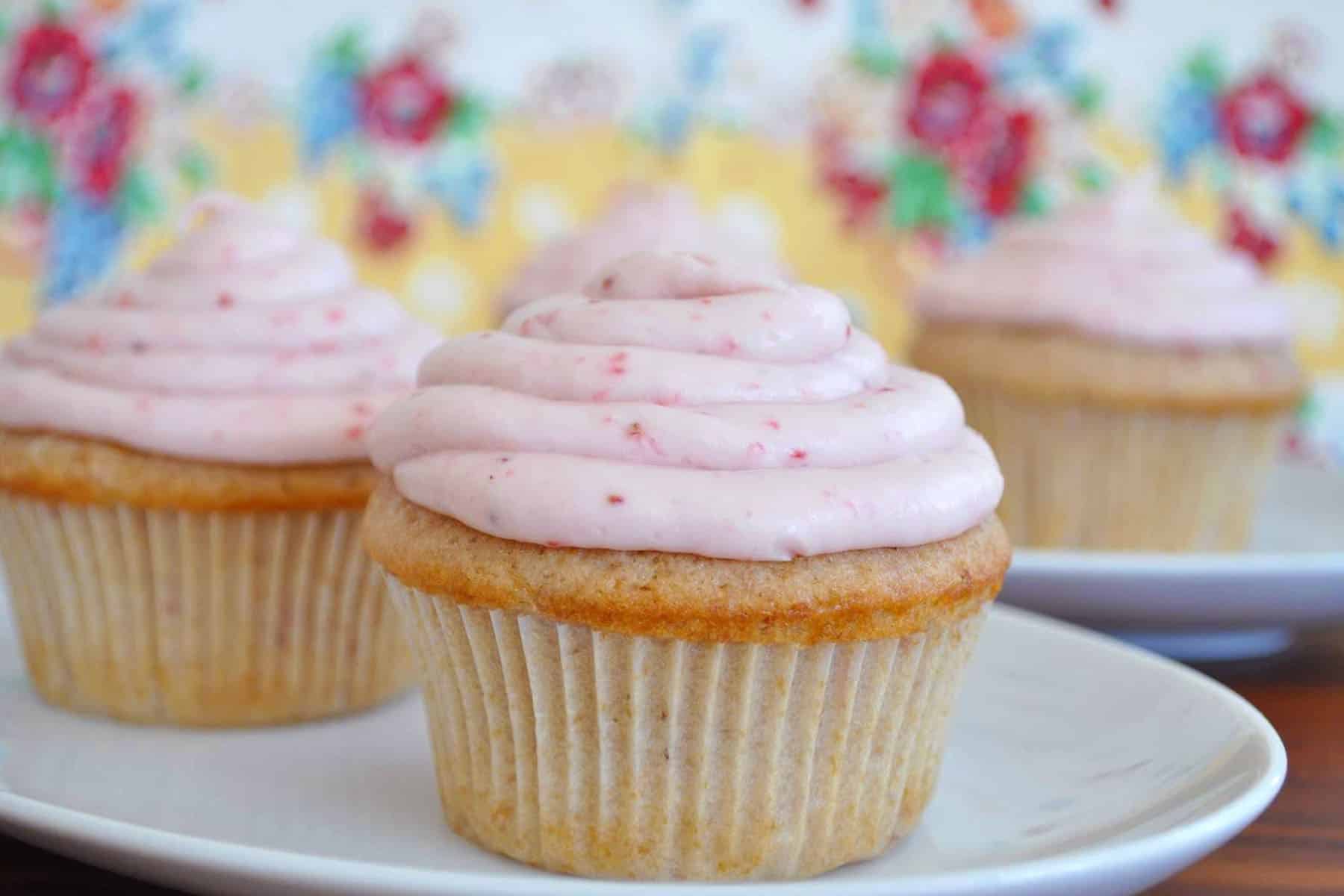 Three vanilla strawberry cupcakes with light pink frosting are displayed on white plates, with a colorful floral-patterned background.