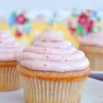 A vanilla cupcake with pink frosting sits on a white plate, reminiscent of classic strawberry cupcakes, with more cupcakes and a floral-patterned background visible behind it.