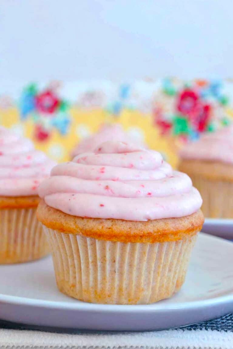 A vanilla cupcake with pink frosting sits on a white plate, reminiscent of classic strawberry cupcakes, with more cupcakes and a floral-patterned background visible behind it.