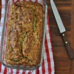 A loaf of banana bread in a glass baking dish sits on a red and white striped towel next to a serrated knife with a wooden handle on a wooden surface.