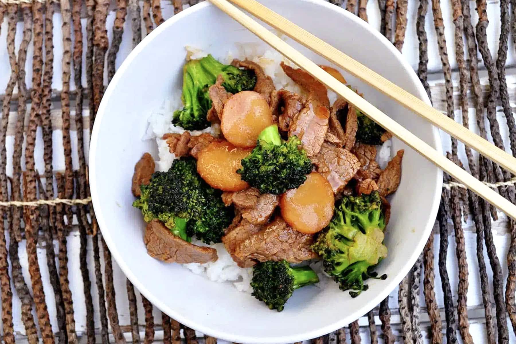 A bowl of Broccoli Beef stir-fry with sliced carrots and white rice, topped with chopsticks, on a woven placemat.