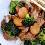 A bowl of white rice topped with savory Broccoli Beef, featuring tender beef slices, crisp broccoli florets, and sliced carrots, with chopsticks resting on the rim.