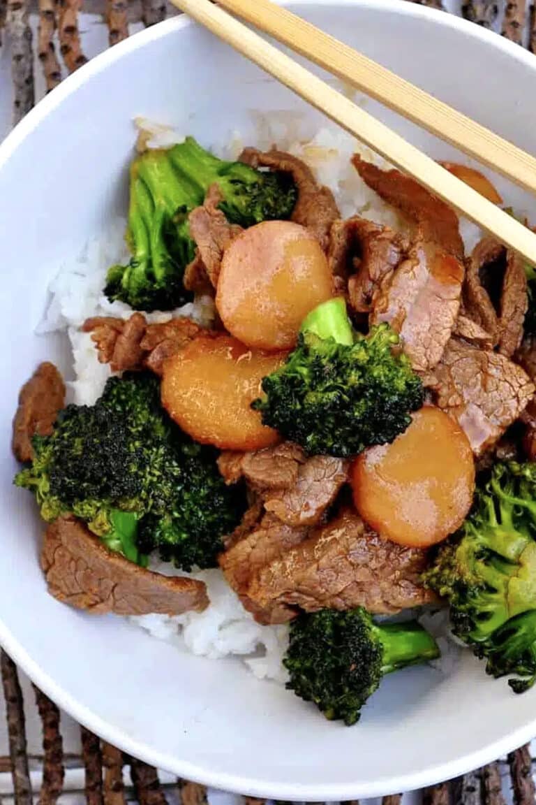 A bowl of white rice topped with savory Broccoli Beef, featuring tender beef slices, crisp broccoli florets, and sliced carrots, with chopsticks resting on the rim.
