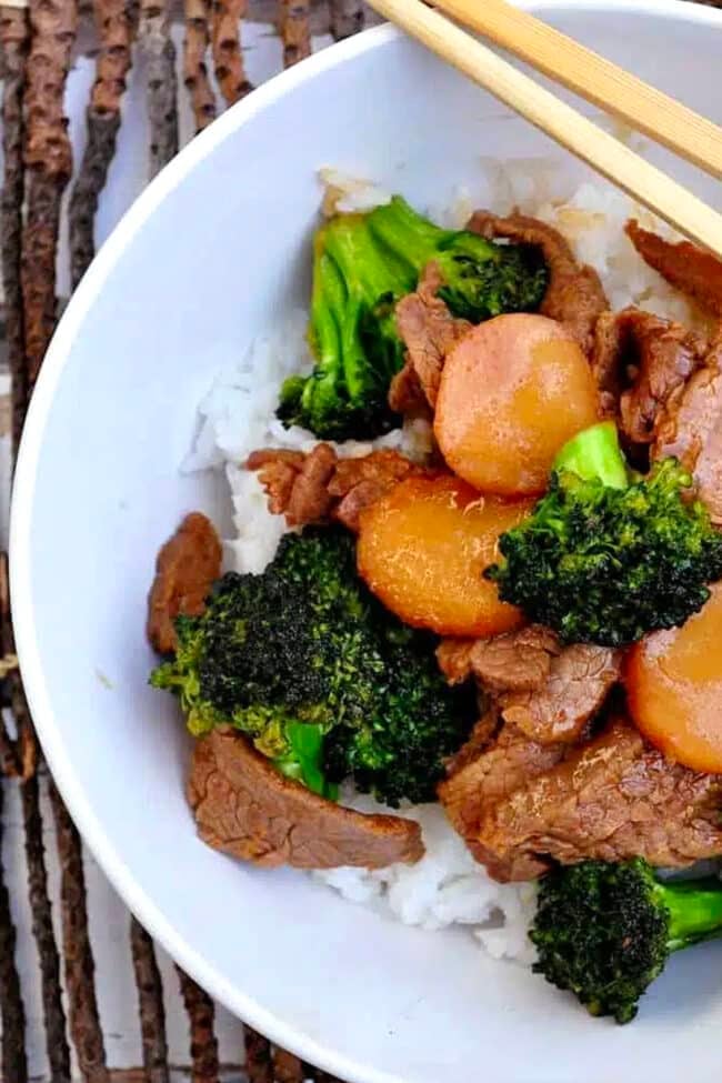 A bowl of fluffy white rice is topped with tender Broccoli Beef, featuring savory cooked beef slices, crisp broccoli florets, and sliced carrots, with wooden chopsticks resting on the bowl’s edge.