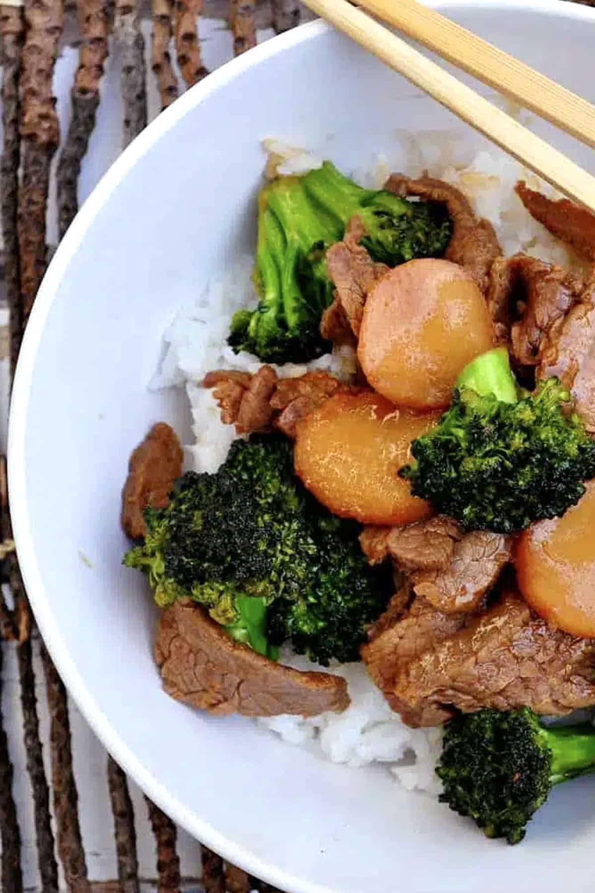 A bowl of fluffy white rice is topped with tender Broccoli Beef, featuring savory cooked beef slices, crisp broccoli florets, and sliced carrots, with wooden chopsticks resting on the bowl’s edge.