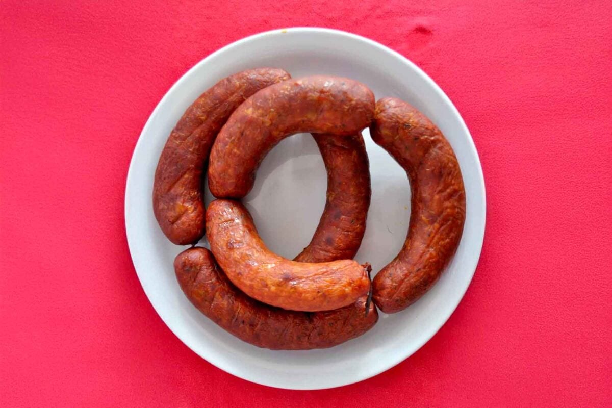 A white plate with four links of homemade linguica arranged in a circular pattern, placed on a bright pink surface.