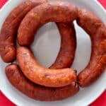 A white plate with four curved, browned homemade linguica sausages arranged in a circle on a bright red tablecloth.