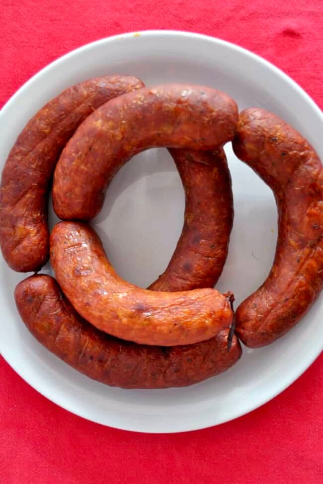 A white plate with four curved, browned homemade linguica sausages arranged in a circle on a bright red tablecloth.