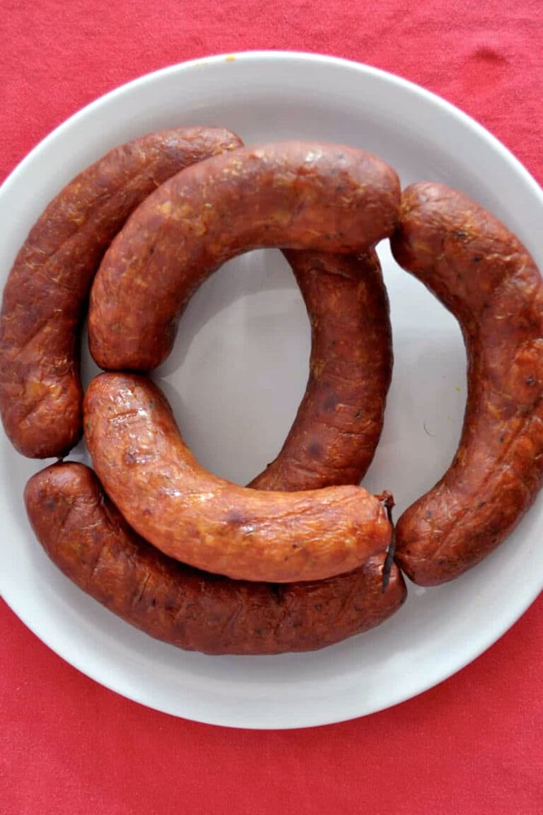 A white plate with four curved, browned homemade linguica sausages arranged in a circle on a bright red tablecloth.