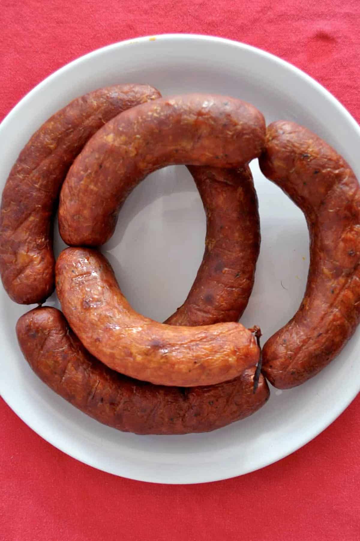 A white plate with four curved, browned homemade linguica sausages arranged in a circle on a bright red tablecloth.