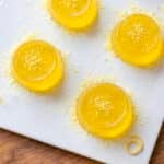 Four yellow gelatin rounds topped with yellow powder on a square white plate, with a small bowl of yellow candies nearby.
