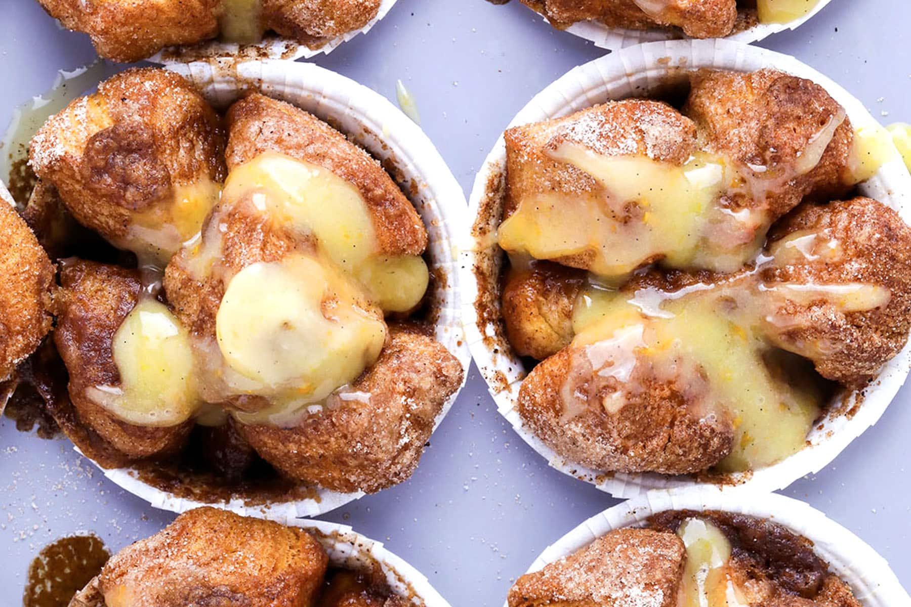 Close-up of several monkey bread muffins in muffin cups, coated in cinnamon sugar and topped with a thick drizzle of yellowish glaze, set against a light purple background.