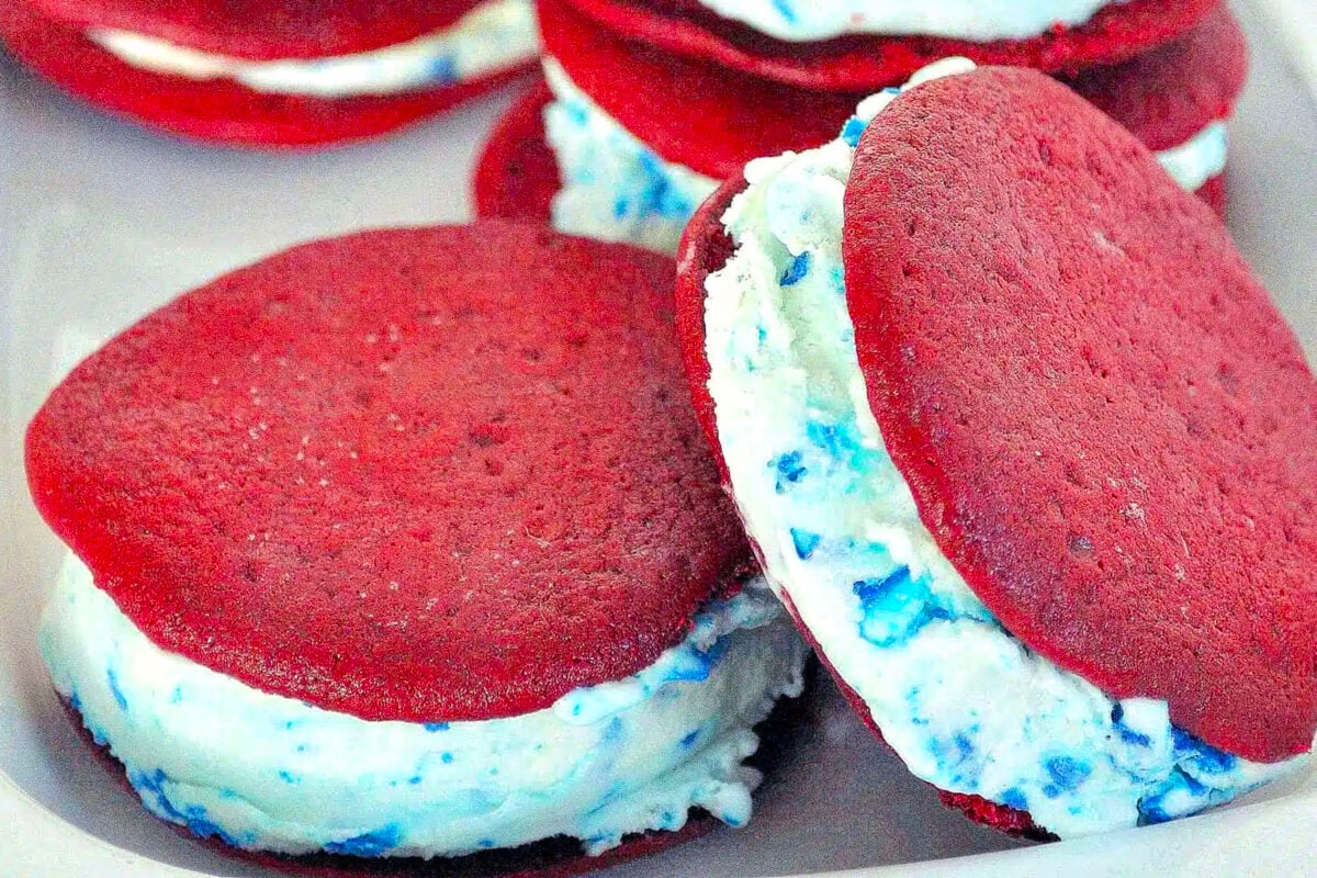 Red velvet ice cream sandwiches filled with white and blue speckled cream are stacked on a tray, showcasing their vibrant red color and creamy centers.