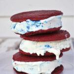 Three delicious ice cream sandwiches are stacked on a white plate, filled with vanilla ice cream containing colorful blue and red sprinkles. The red velvet cookies sandwich the melting ice cream, oozing slightly at the edges.