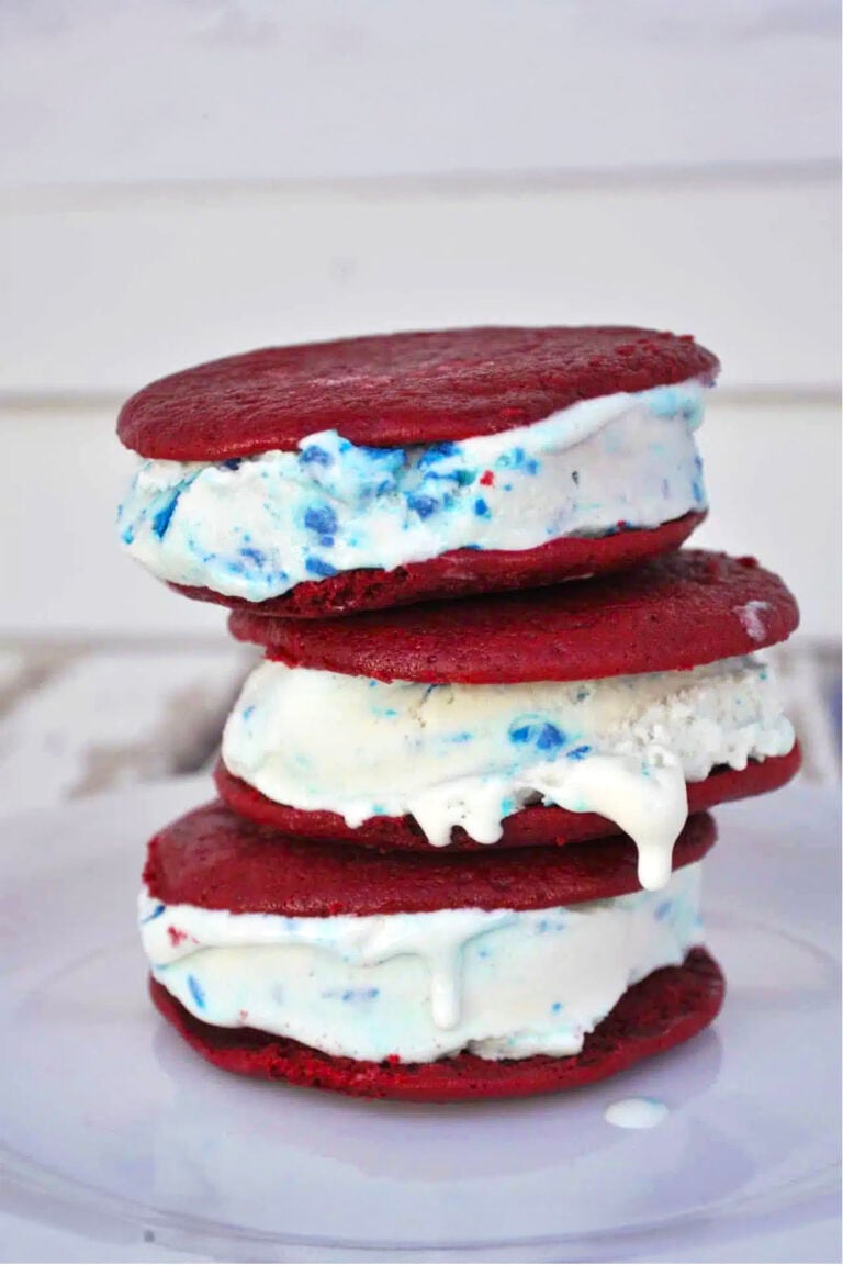 Three delicious ice cream sandwiches are stacked on a white plate, filled with vanilla ice cream containing colorful blue and red sprinkles. The red velvet cookies sandwich the melting ice cream, oozing slightly at the edges.