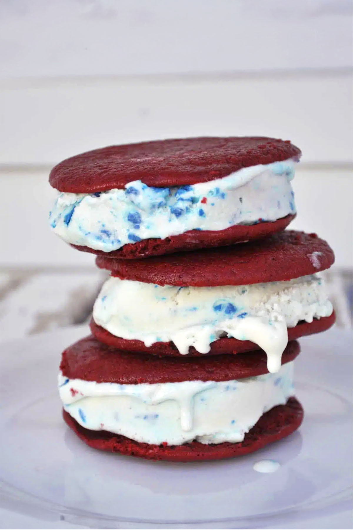 Three delicious ice cream sandwiches are stacked on a white plate, filled with vanilla ice cream containing colorful blue and red sprinkles. The red velvet cookies sandwich the melting ice cream, oozing slightly at the edges.