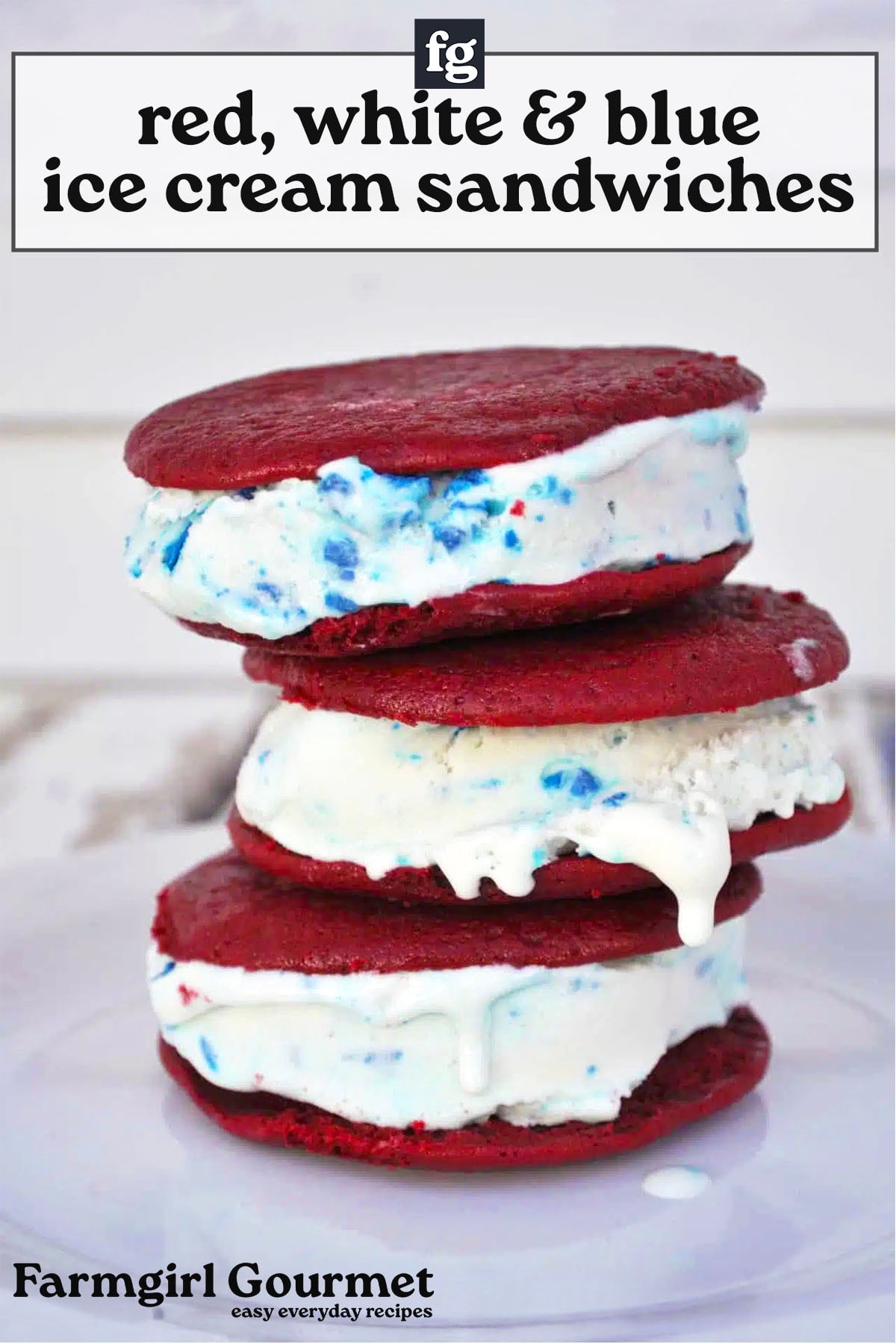 Three ice cream sandwiches made with red velvet cookies, white ice cream, and blue and red candy pieces are stacked on a plate. Text above them reads red, white & blue ice cream sandwiches.