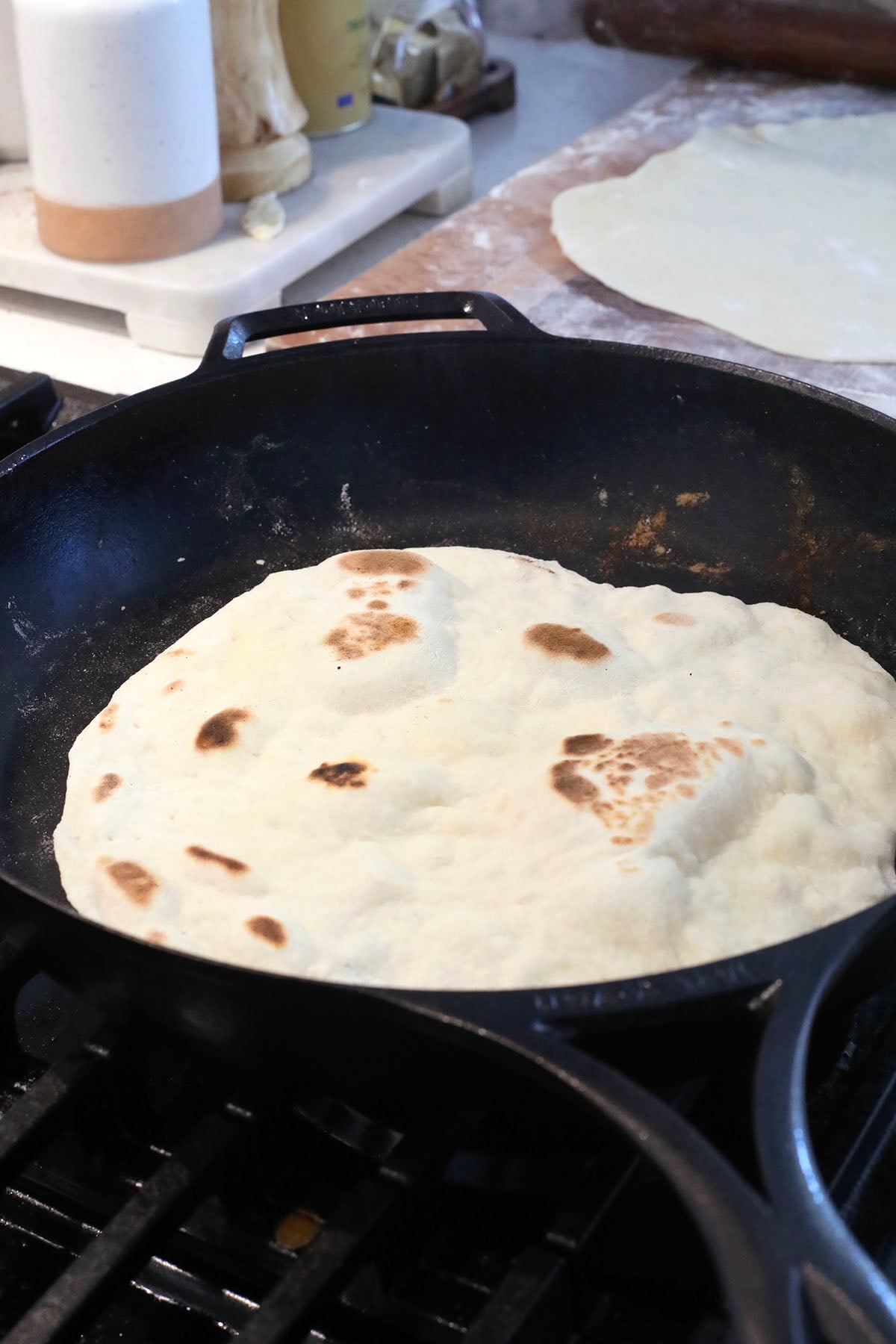 A clear glass bowl filled with ingredients for homemade flour tortillas—flour, baking powder, salt, and sugar—sits on a marble countertop next to a measuring cup containing oil.