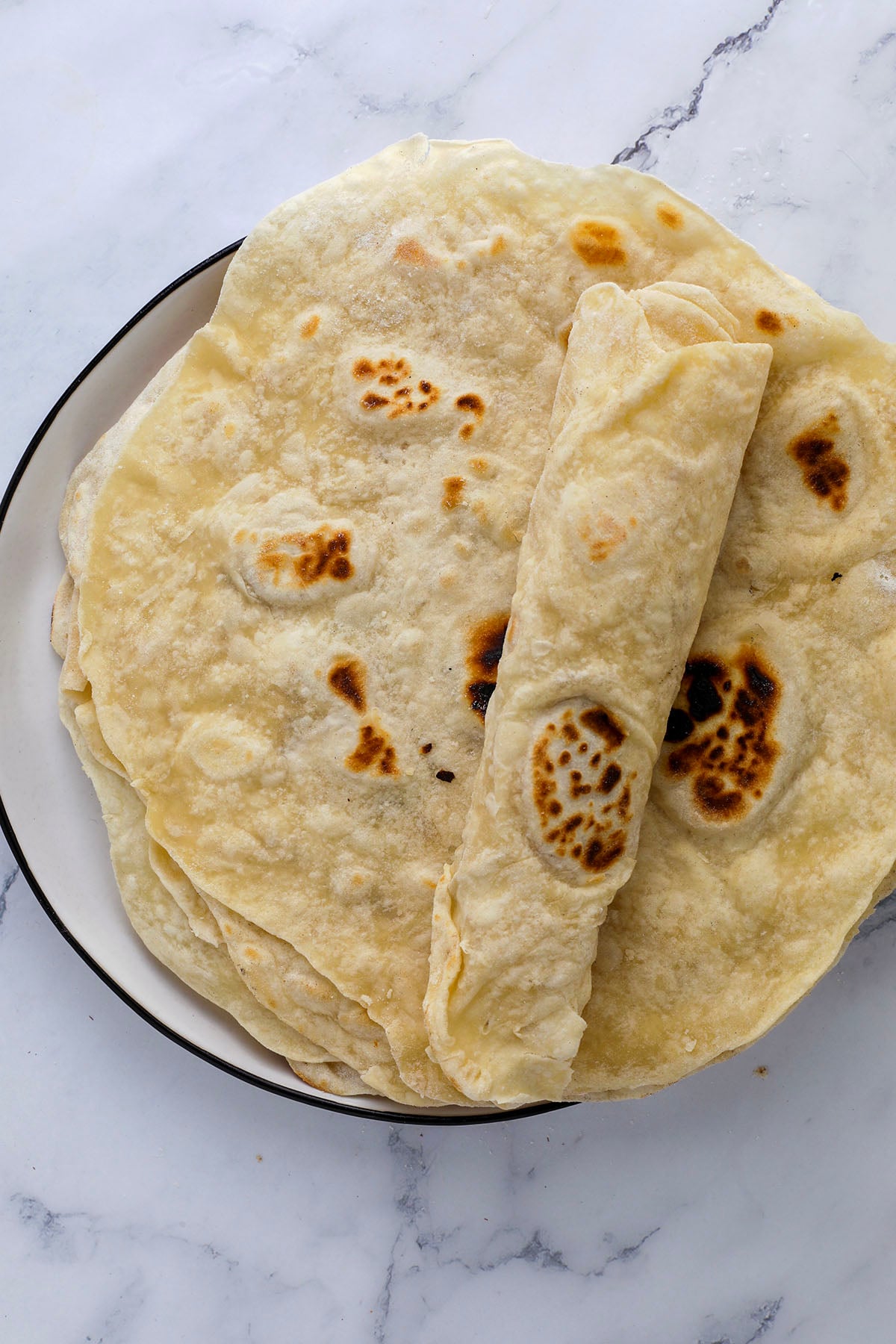 A clear glass bowl filled with ingredients for homemade flour tortillas—flour, baking powder, salt, and sugar—sits on a marble countertop next to a measuring cup containing oil.