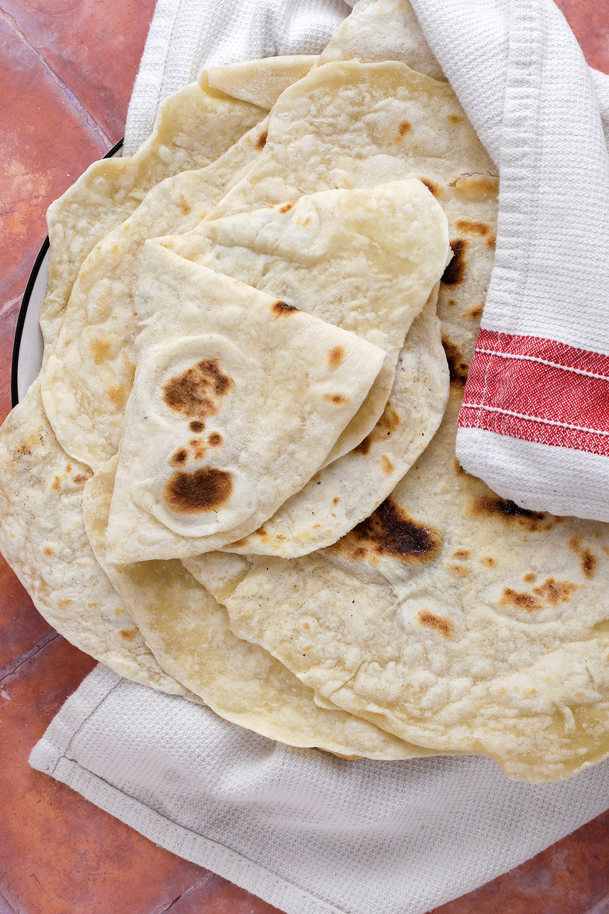 A plate of homemade flour tortillas, partially covered with a white kitchen towel featuring a red stripe, sits on a reddish-brown surface. The golden flatbreads with toasted spots showcase the results of a classic flour tortilla recipe.