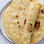 A plate of homemade flour tortillas, with one rolled up and placed on a stack, sits on a white marble surface—a perfect showcase for an easy flour tortilla recipe.