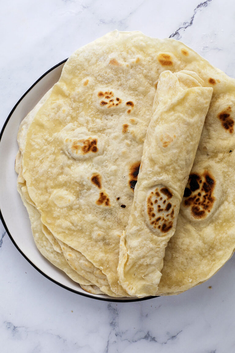 A plate of homemade flour tortillas, with one rolled up and placed on a stack, sits on a white marble surface—a perfect showcase for an easy flour tortilla recipe.