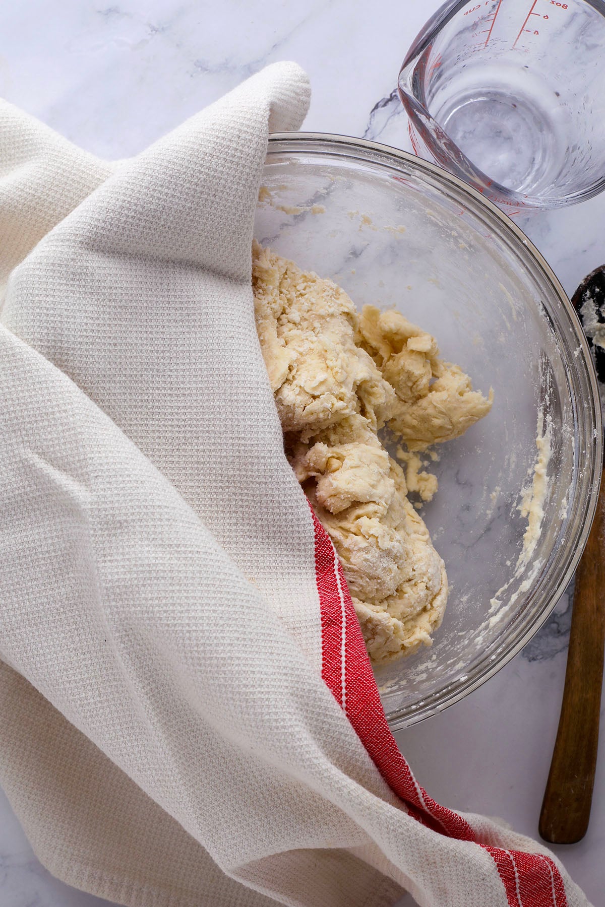 A clear glass bowl filled with ingredients for homemade flour tortillas—flour, baking powder, salt, and sugar—sits on a marble countertop next to a measuring cup containing oil.