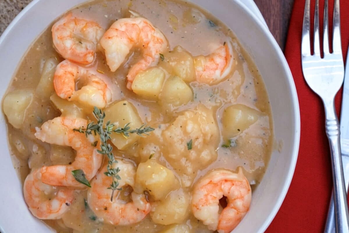 A bowl of Cajun shrimp stew with tender potatoes, garnished with fresh thyme, placed next to a fork and red napkin on a wooden surface.