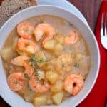A bowl of creamy Cajun shrimp and potato stew garnished with herbs, served with multigrain bread on a white plate, next to a red napkin, fork, and knife on a wooden table.
