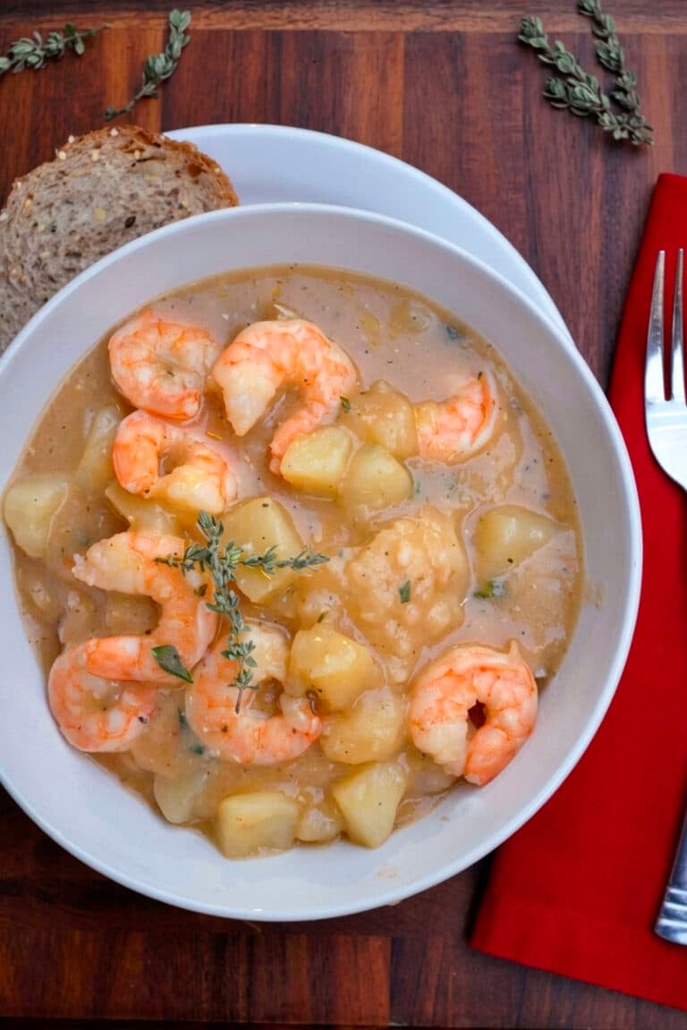 A bowl of creamy Cajun shrimp and potato stew garnished with herbs, served with multigrain bread on a white plate, next to a red napkin, fork, and knife on a wooden table.