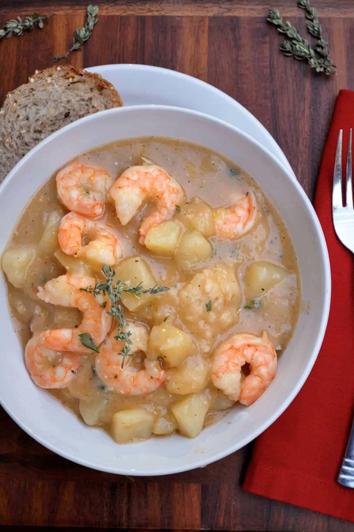A bowl of creamy Cajun shrimp and potato stew garnished with herbs, served with multigrain bread on a white plate, next to a red napkin, fork, and knife on a wooden table.